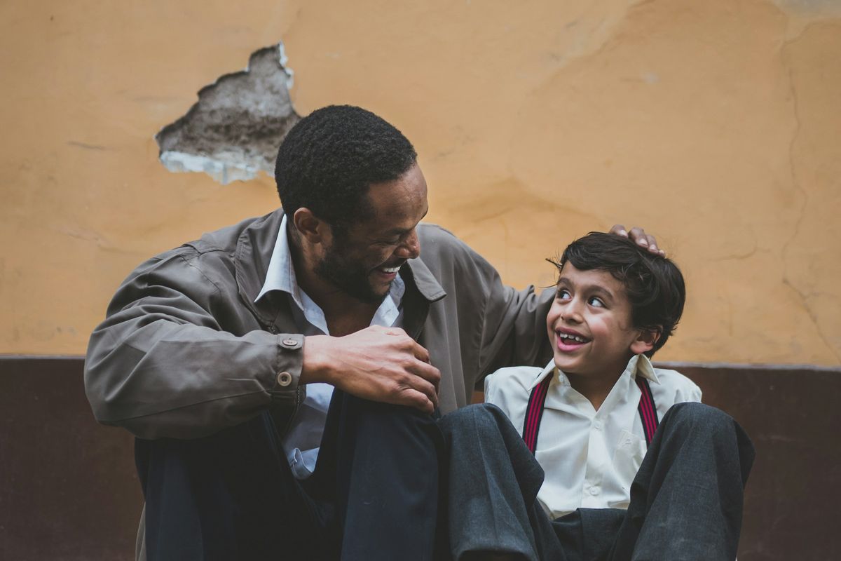 A father and his young son sit together on a step, smiling and talking.