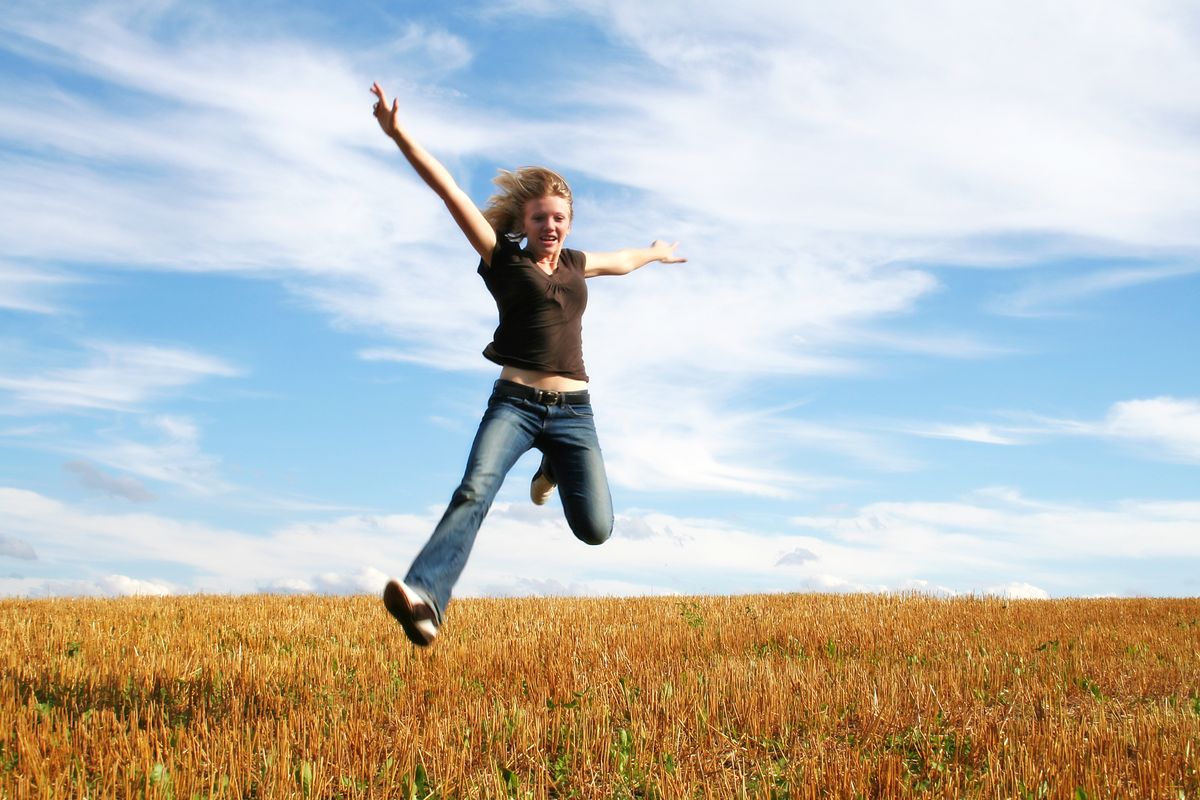 A woman leaps for joy in a wheat field under a wide blue sky.